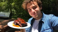 Smiling person outdoors holding a plate of barbecued ribs with sauce, picnic table in background.